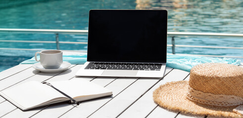 Alternative workplace at resort poolside. Laptop, notepad, cup of coffee and hat on white table isolated on blue background of pool water. Blank computer screen, copy space for text or design. Banner