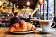 In the foreground, a delicious fresh croissant next to a cup of cappuccino with a blurred cafe background create a cozy morning atmosphere