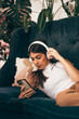A young dark-haired woman in casual clothes wearing headphones,using a smartphone,lying on the couch.Relaxation,meditation,listening music.