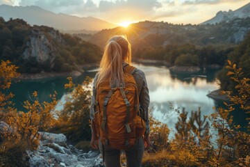 Wall Mural - A woman with a backpack stands at the edge of a cliff, beholding the sunset over a serene mountain lake amidst autumn foliage