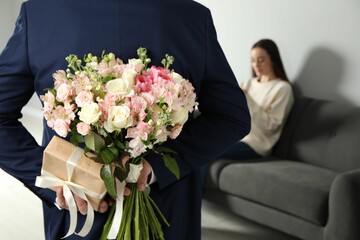 Wall Mural - Man hiding bouquet of flowers and present for his beloved woman indoors, closeup