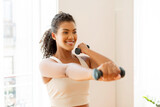 Fototapeta Panele - Cheerful young woman in sportswear holds dumbbells and exercising indoor
