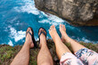 A couple sits on a cliff with their feet dangling over an abyss above the ocean. First person photo.
