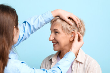 Poster - Young woman putting hearing aid in mature woman's ear on blue background