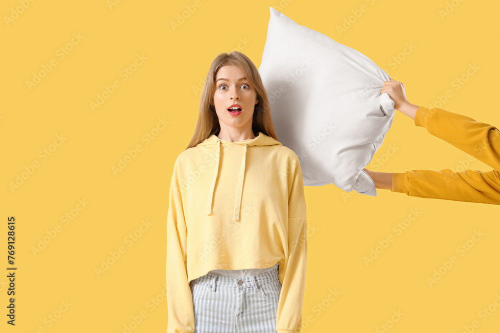 Shocked young woman getting hit with pillow on yellow background