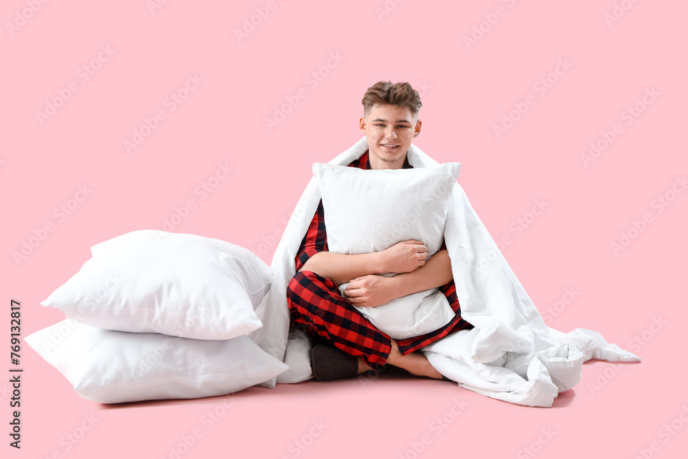 Young man in pajamas with soft pillows and blanket sitting on pink background