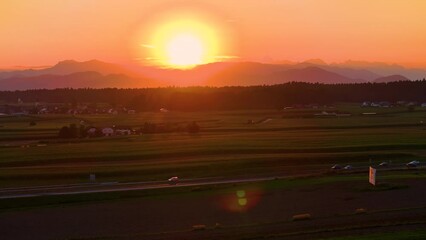 Wall Mural - AERIAL, LENS FLARE: Sunset illuminates the idyllic countryside, with commuters driving along scenic highway. Golden hour evening sun rays shine on the countless cars driving down the rural freeway.