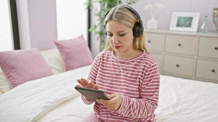 Poster - A young woman wearing headphones focuses on a tablet while sitting on a bed in a cozy, well-decorated bedroom.