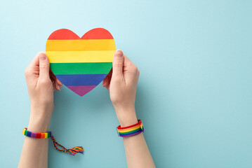 Sticker - Rainbow heart-shaped card held by a woman's hands wearing symbolic bracelet and wristlet in a first-person top view on a pastel blue background with space for text or advertising