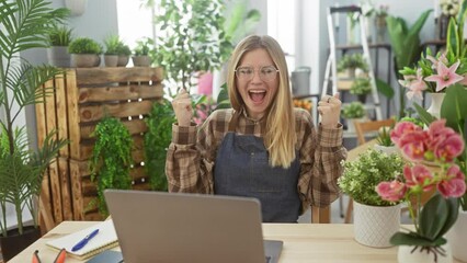 Sticker - A joyful blonde woman wearing glasses celebrates success at her laptop in a vibrant flower shop setting.