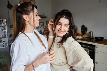 happy young woman holding glass of fresh orange juice and flirting with girlfriend in kitchen, lgbt