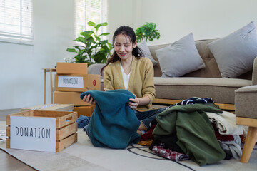 Wall Mural - Asian young woman packing clothes at home, putting on stuff into donate box with second hand clothes. Help poor people. Donation concept