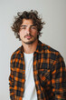 © ChubbyCat - Portrait of a young man with curly hair and stubble wearing a plaid shirt over a white t-shirt, posing in front of a neutral background with a confident gaze.