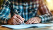Person in a plaid shirt writing notebook with pen, close-up on hands and paper.