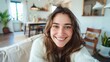 © Z A C - Smiling woman with long brown hair wearing a white sweater sitting in a cozy living room with a wooden floor plants and a window in the background.