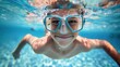 A young boy wearing blue goggles smiling underwater with bubbles around his face in a swimming pool.