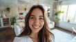 © Z A C - Smiling woman with long brown hair wearing a white top standing in a modern living room with natural light.