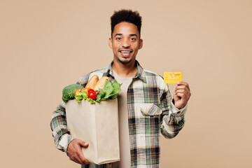 Wall Mural - Young smiling man wears grey shirt hold paper bag for takeaway mock up with food products, credit bank card isolated on plain pastel light beige background. Delivery service from shop or restaurant.