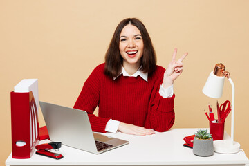 Wall Mural - Young smiling happy employee business woman she wear red sweater shirt sit work at office desk with pc laptop showing victory sign isolated on plain pastel beige background Achievement career concept