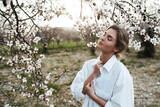 Fototapeta  - Beautiful young woman wearing a white shirt, elegantly posing near the blossoming almond trees