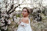 Fototapeta Zachód słońca - Beautiful young woman wearing a white shirt, elegantly posing near the blossoming almond trees