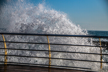 Wall Mural - Huge waves roll onto a wooden pier with railings against the backdrop of a clear blue sky and sea horizon	
