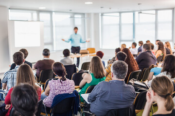 Wall Mural - Business and entrepreneurship symposium. Speaker giving a talk at business meeting. Audience in conference hall. Rear view of unrecognized participant in audience.