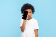 © khosrork - Portrait of funny playful little boy with curly hair covering his eye with smartphone with empty display for advertisement. Indoor studio shot isolated on blue background.