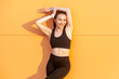 © khosrork - Portrait of pretty joyful positive attractive woman wearing black sportswear standing with raised arms looking at camera showingher slim sporty body. Outdoor shot against orange wall.