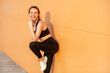 © khosrork - Portrait of attractive smiling woman wearing black sportswear looking at camera with satisfied face finishing training resting after workout. Outdoor shot against orange wall.