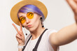 Portrait of funny cool woman blogger with violet hair in sunglasses and hat making selfie POV showing v sign grimacing. Indoor studio shot isolated on white background.