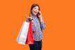 Portrait of cute charming woman with brown hair holding shopping bags and talking phone, boasting purchases, wearing checkered shirt. Indoor studio shot isolated on orange background