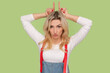 Portrait of angry aggressive adult blond woman making devil cow horns, being ready to attack, arguing with somebody, wearing denim overalls. Indoor studio shot isolated on light green background