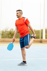 Wall Mural - An adult man between 30 and 40 years old is playing a game of pickleball outdoors.The young man dressed in sportswear is stretching one leg after training.Pickleball concept.