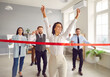 © Studio Romantic - Happy woman employee running to the red ribbon with raised arms celebrating crossing finishing line in office with people colleagues in background. Business competition and teamwork concept.