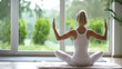 Woman practicing yoga in a seated position with raised arms indoors facing a window with a view of greenery outside.