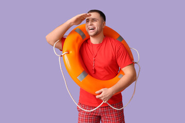 Wall Mural - Happy young lifeguard with whistle and lifebuoy looking at somewhere on lilac background