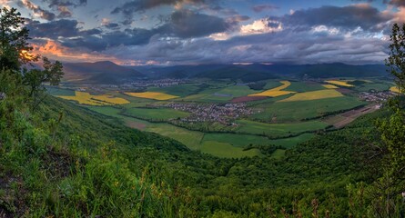 Poster - View from the hill of a spring landscape with fields of canola, grain and forests. Small villages in the valley, hilly landscape with dramatic sunset sky