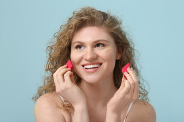 Wall Mural - Beautiful young woman with foundation on her face holding makeup sponges against blue background