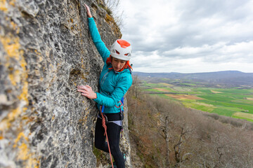 A woman is climbing a rock wall wearing a helmet. Concept of adventure and excitement, as the woman is taking on a challenging physical activity. The cloudy sky adds a dramatic backdrop to the scene