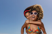 An Adult And A Child Share A Joyous Hug And Laughter Against A Clear Blue Sky On A Sunny Beach Day, Showcasing Warmth And Happiness