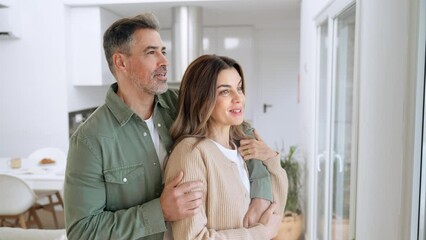 Wall Mural - Happy middle aged mature couple hugging looking away relaxing standing at home. Relaxed serene older man and woman in love bonding together embracing in house living room looking at window.