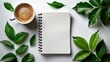 © Surachetsh - flat lay photograph showcasing blank white spiral notebook resting on table adorned with coffee cups and verdant green leaves, against minimalist white background.