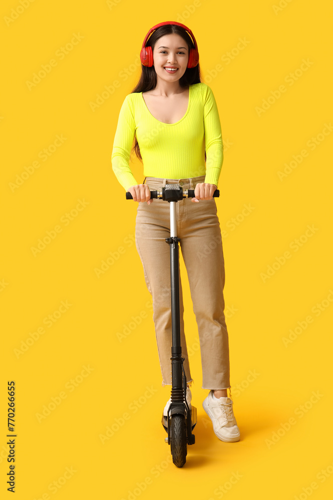 Beautiful young Asian woman in headphones with modern electric kick scooter on yellow background