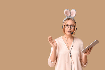 Surprised adult female operator in bunny ears with headset and tablet computer on beige background. Easter celebration