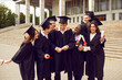 © Studio Romantic - Group of a smiling happy multiracial international graduates students hugging and having fun in a university graduate gown holding diploma outdoor. Education and graduation concept.