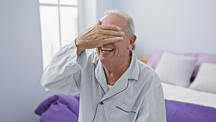 Wall Mural - Cheerful senior man in pyjamas covers his eyes playfully, sitting on his bedroom bed, embracing surprise with a radiant smile