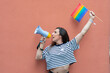 transgender LGBT hispanic celebrating rainbow flag and megaphone announcing equality isolated on nude colored background.