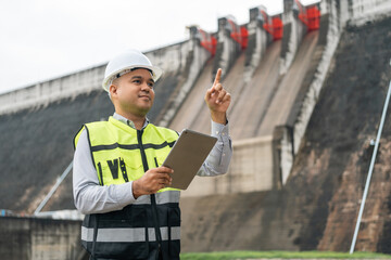 Wall Mural - Professional asian maintenance engineer man with safety helmet in construction site dam with hydroelectric power plant and irrigation. Manager engineer man working with tablet at project big building.