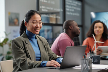 Leinwandbilder - Happy young Asian female employee in formalwear and eyeglasses networking by desk with windmill model against two colleagues